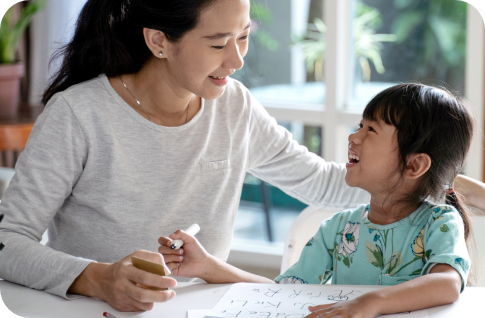 Mother and daughter sit at a table learning, with paper, letters, and pencils. Both smile warmly, enjoying the educational activity at home.