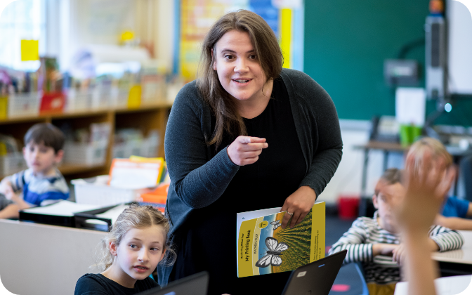 A smiling teacher leans toward students with a book in hand, engaging them. Children sit at desks with laptops, one raising a hand, creating a lively classroom scene.