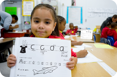 Young girl in a classroom proudly holds up a handwriting practice sheet featuring the LWT workbook. Other children are working at a table in the background.
