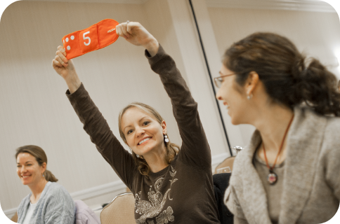 Participant in a classroom training session holds up an orange card with the number 5 while seated among other attendees in a meeting room.