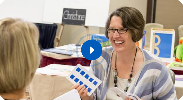 Two women sit at a table; one smiles while holding a card with blue squares. Educational materials and a whiteboard are in the background.